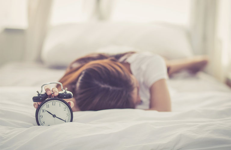 Une femme est allongée sur le ventre sur un lit et tient un réveil dans sa main étendue.